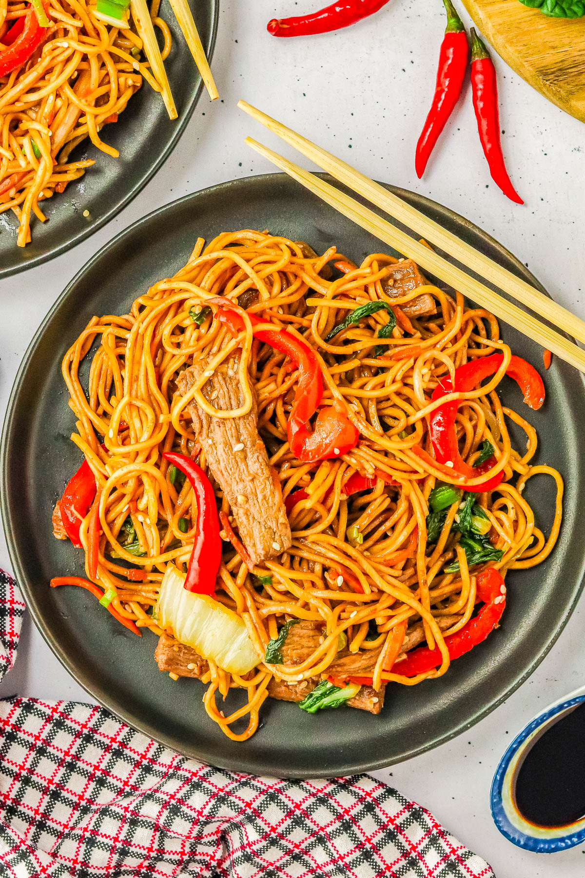 A plate of stir-fried noodles with vegetables and meat, accompanied by chopsticks. Red chili peppers and a checkered cloth are visible in the background.