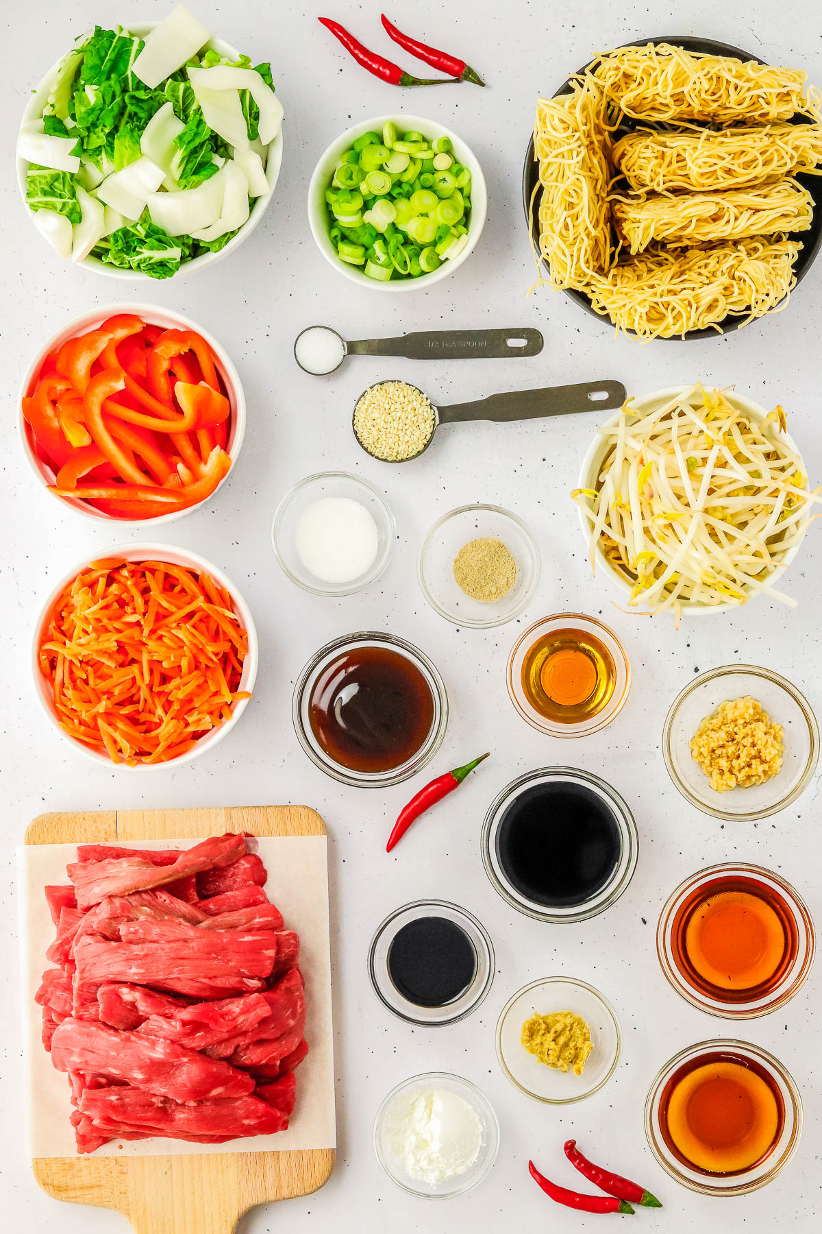 Various ingredients for a stir-fry, including sliced beef, red bell peppers, green onions, bok choy, carrots, sauces, spices, noodles, bean sprouts, and fresh red chilies, arranged on a white surface.