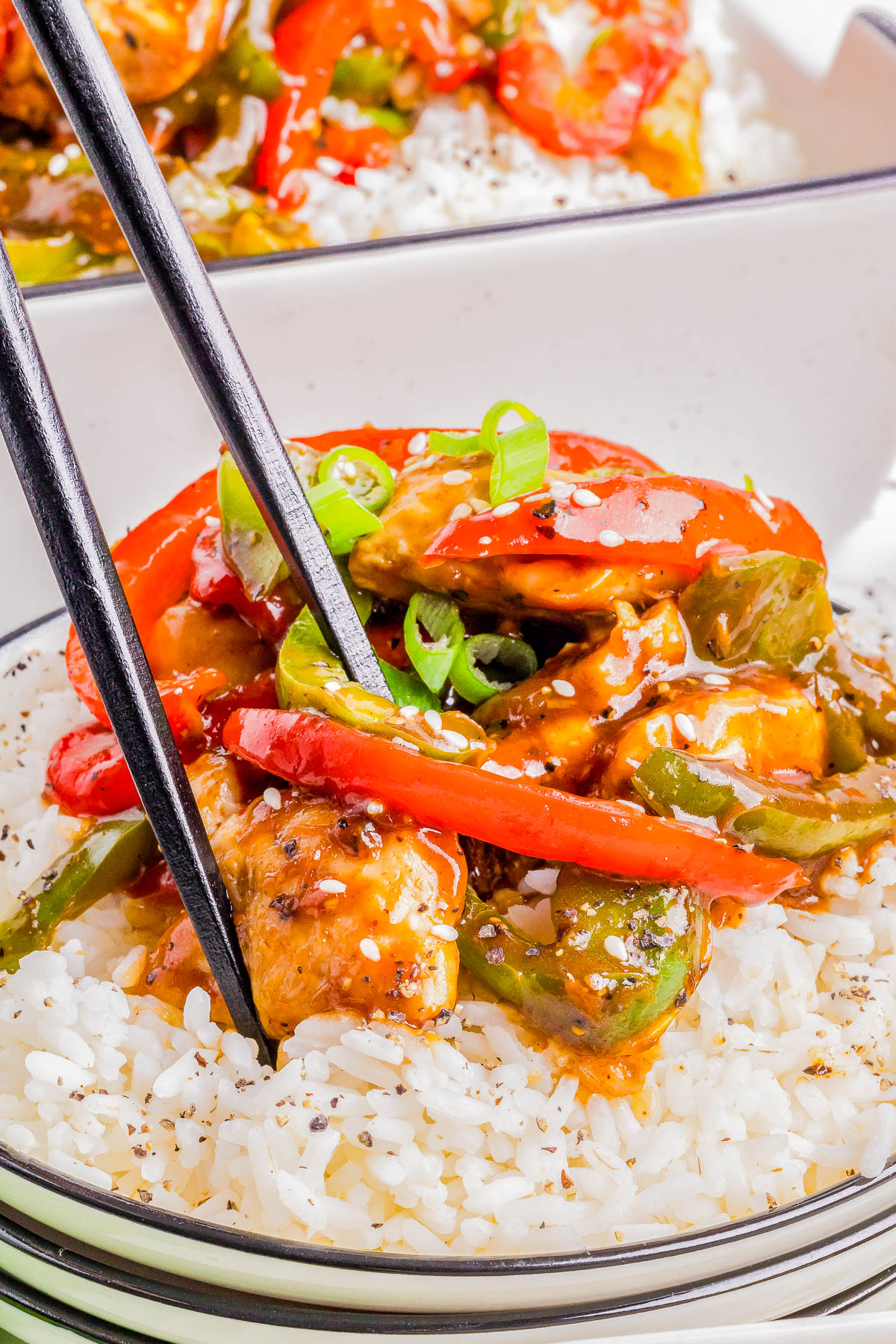 A bowl of white rice topped with stir-fried chicken, red and green bell peppers, and green onions, with a pair of black chopsticks picking up food.