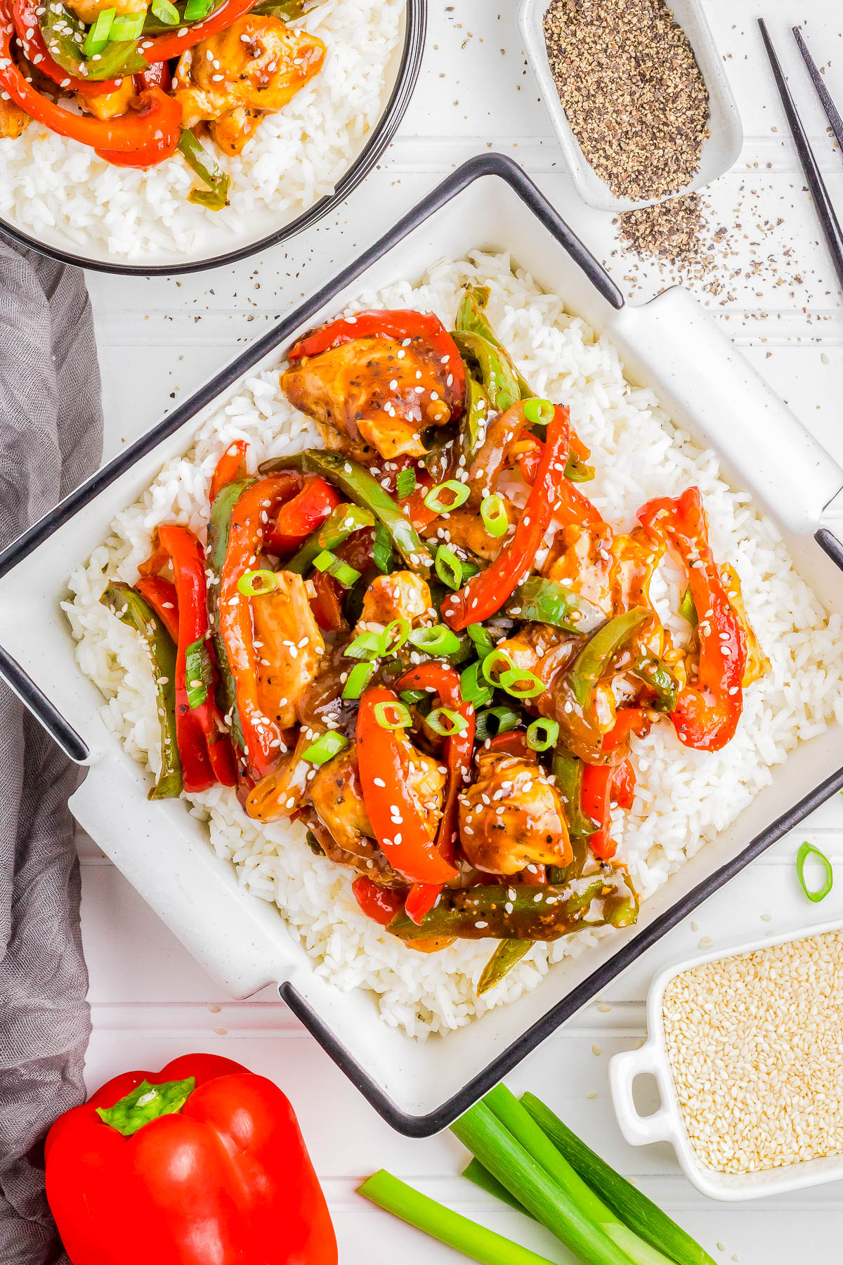 A square dish filled with white rice is topped with stir-fried chicken, red and green bell peppers, and garnished with sesame seeds and green onions. A full red bell pepper and green onions are beside the dish.