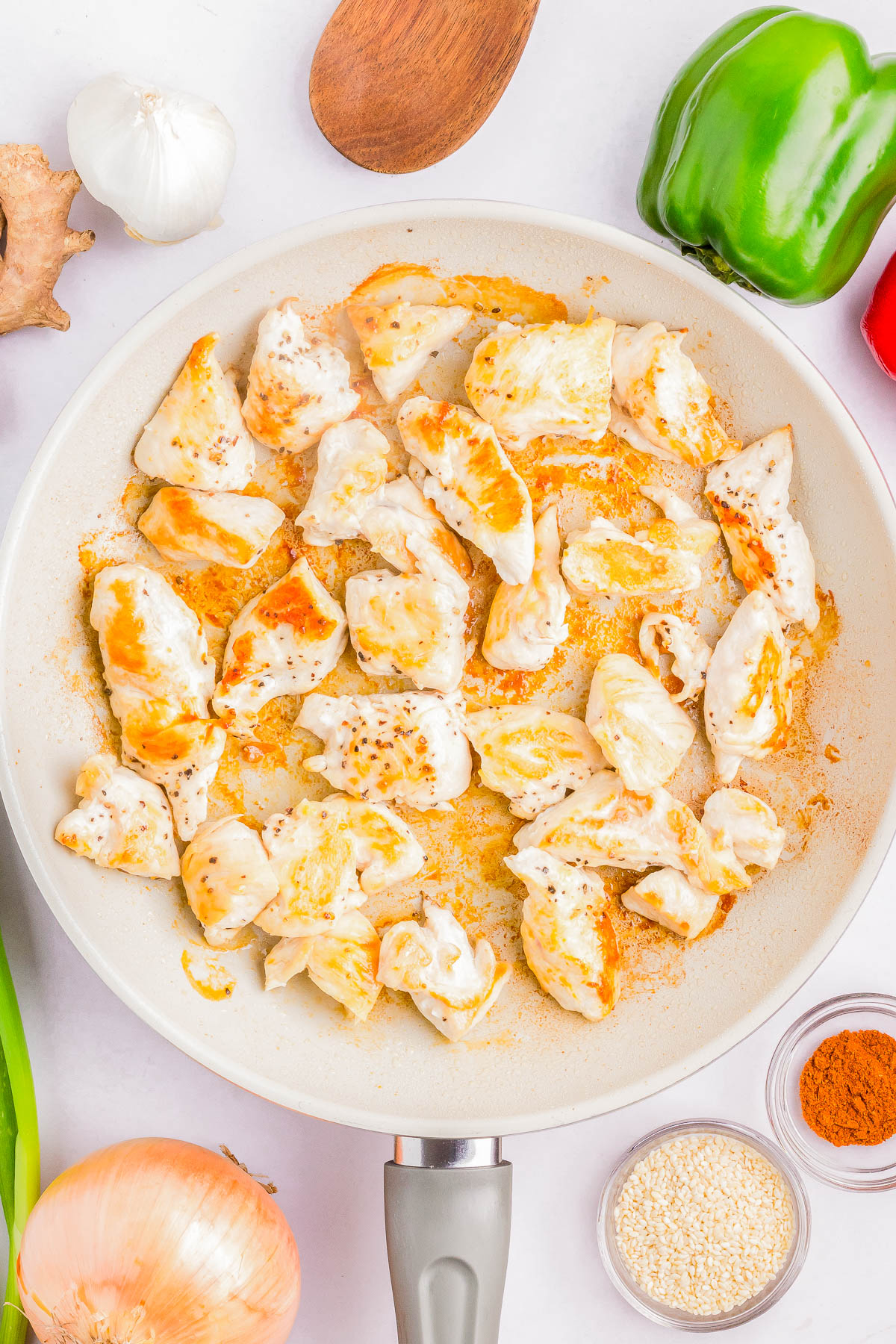 A pan with browned chunks of chicken, surrounded by a green bell pepper, garlic, ginger, a red bell pepper, an onion, and spices.