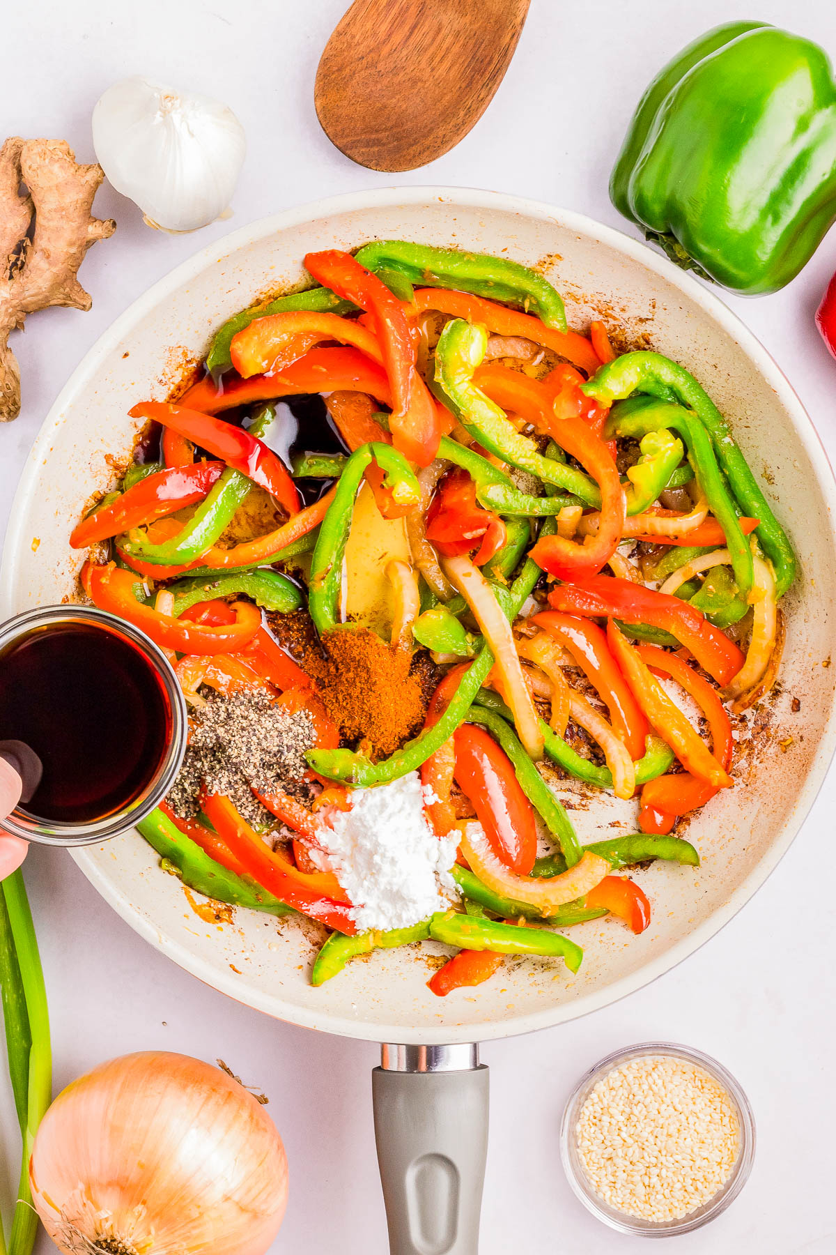 A pan with sliced red and green bell peppers, onions, and seasoning powder. A hand pours dark liquid into the pan. Nearby are garlic, ginger, a wooden spoon, and other ingredients.