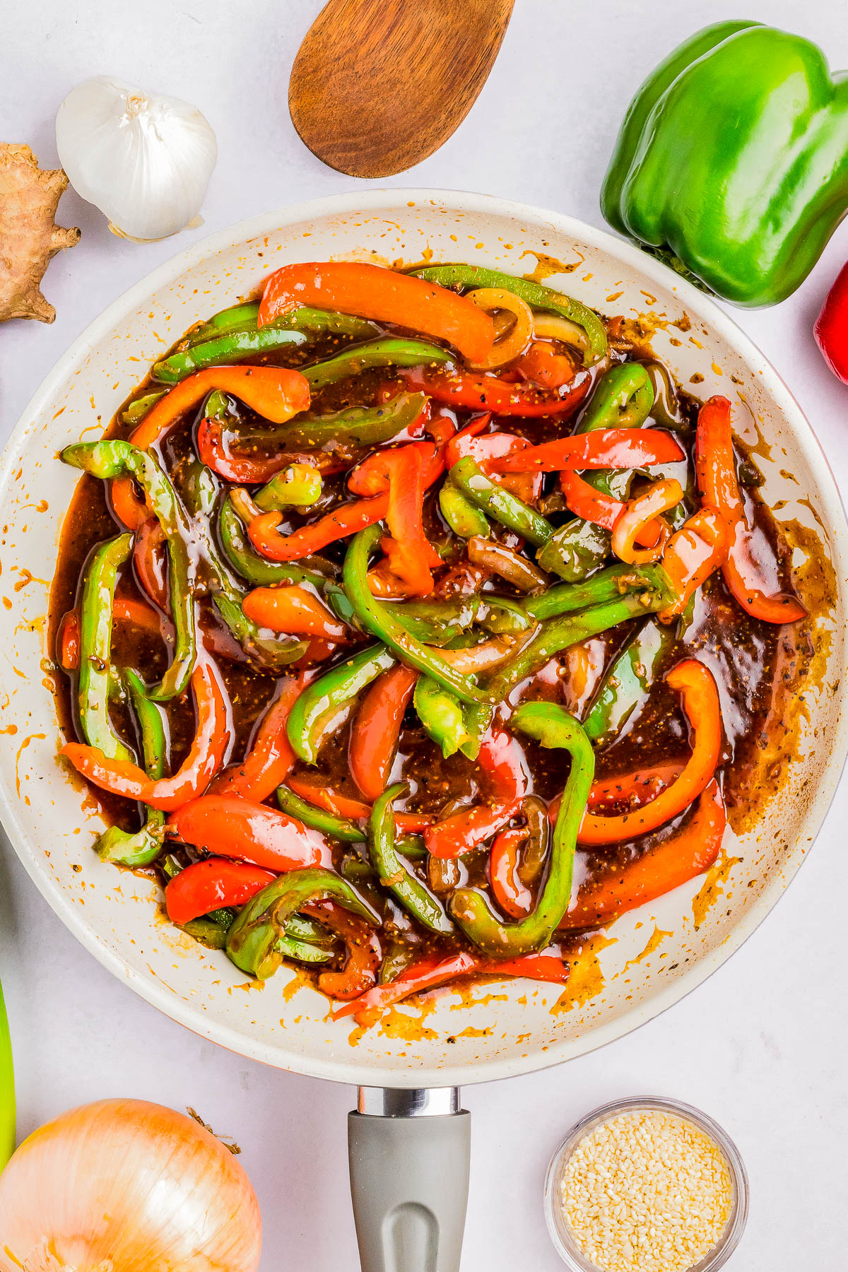 A skillet with sliced red and green bell peppers in a brown sauce, surrounded by whole vegetables and a wooden spoon.