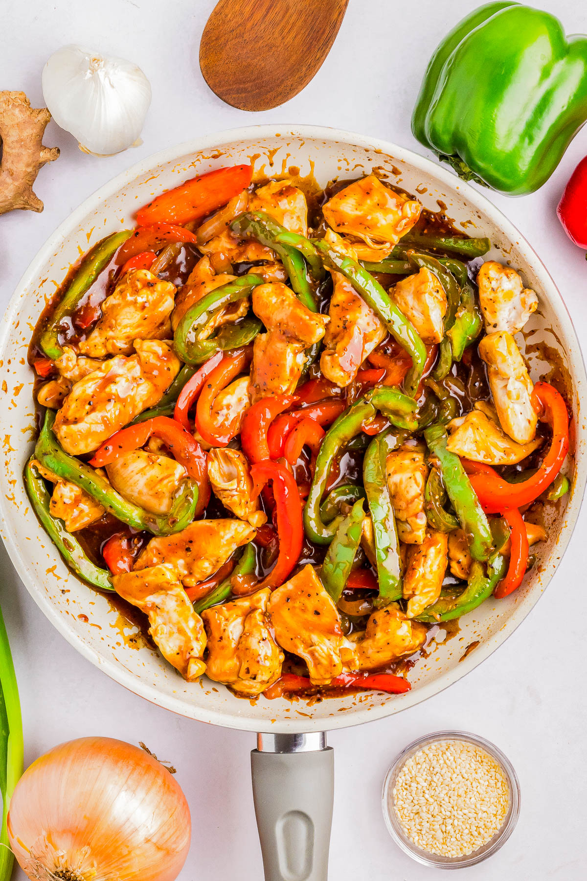 A skillet filled with cooked chicken strips, red and green bell peppers, in a brown sauce. Garlic, ginger, onion, green onion, sesame seeds, and wooden utensil surround the skillet.