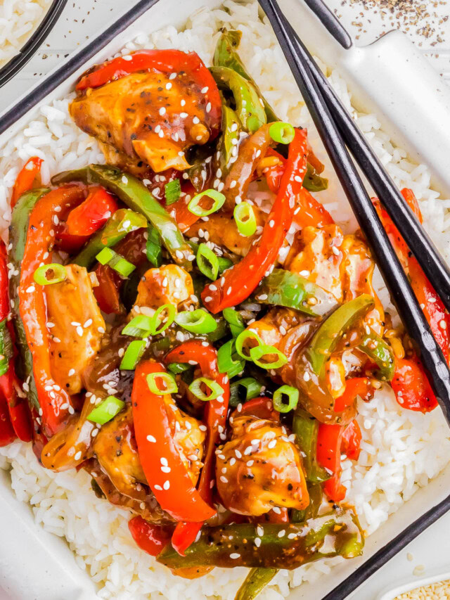 A close-up of a plate of stir-fry with chicken, bell peppers, and green onions served over white rice. Chopsticks rest on the plate.