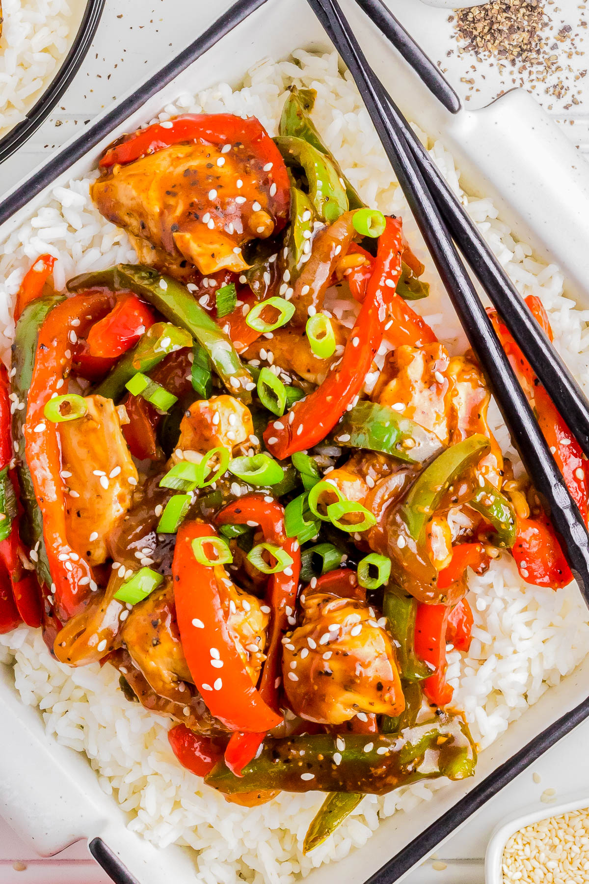 A close-up of a plate of stir-fry with chicken, bell peppers, and green onions served over white rice. Chopsticks rest on the plate.