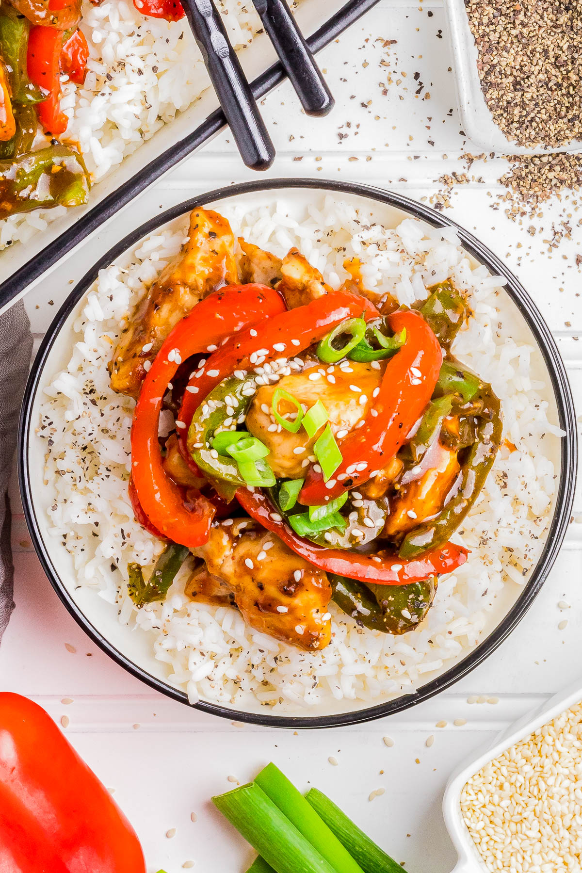 A bowl of white rice topped with stir-fried chicken, bell peppers, and green onions, garnished with sesame seeds. Black chopsticks rest beside the dish.