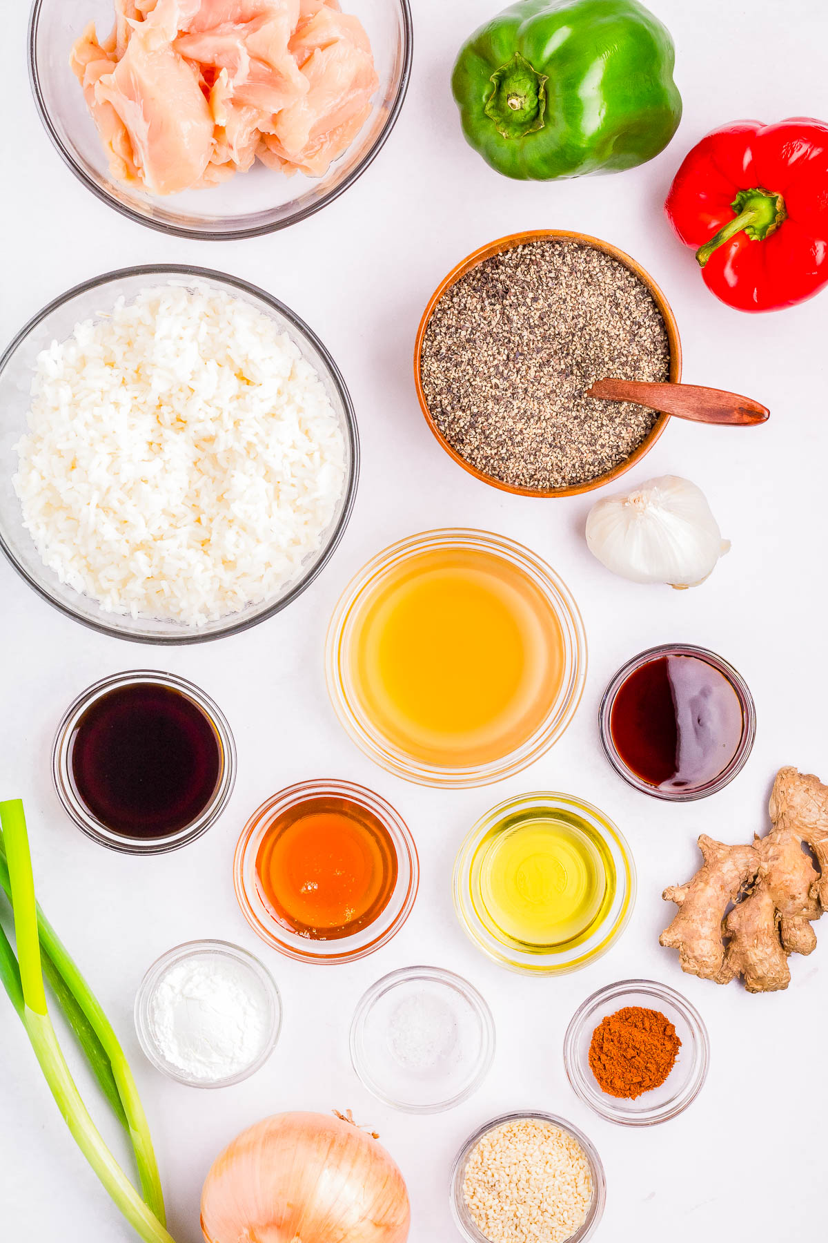 Top-down view of various ingredients organized on a white surface, including raw chicken, bell peppers, rice, sauces, garlic, ginger, spices, and cooking oils.