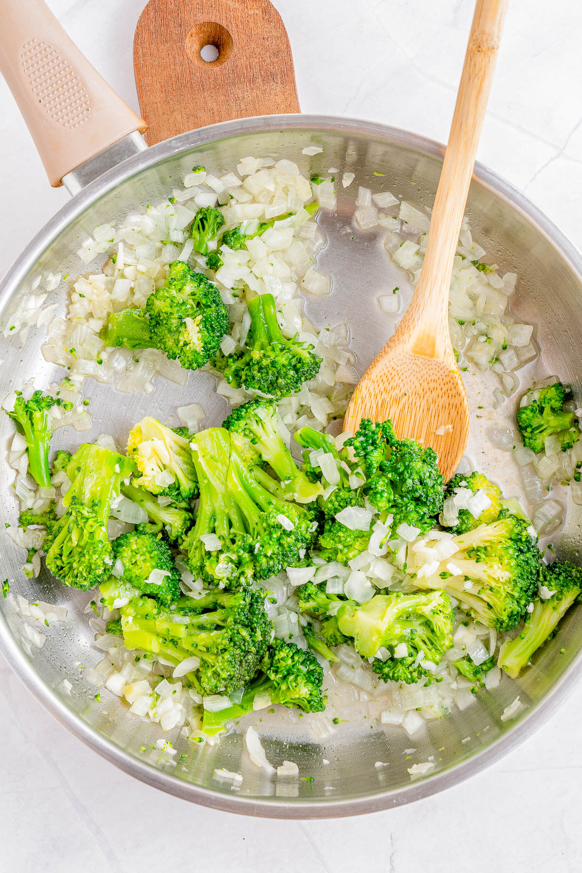 A skillet with cooked broccoli florets and chopped onions being stirred with a wooden spoon.