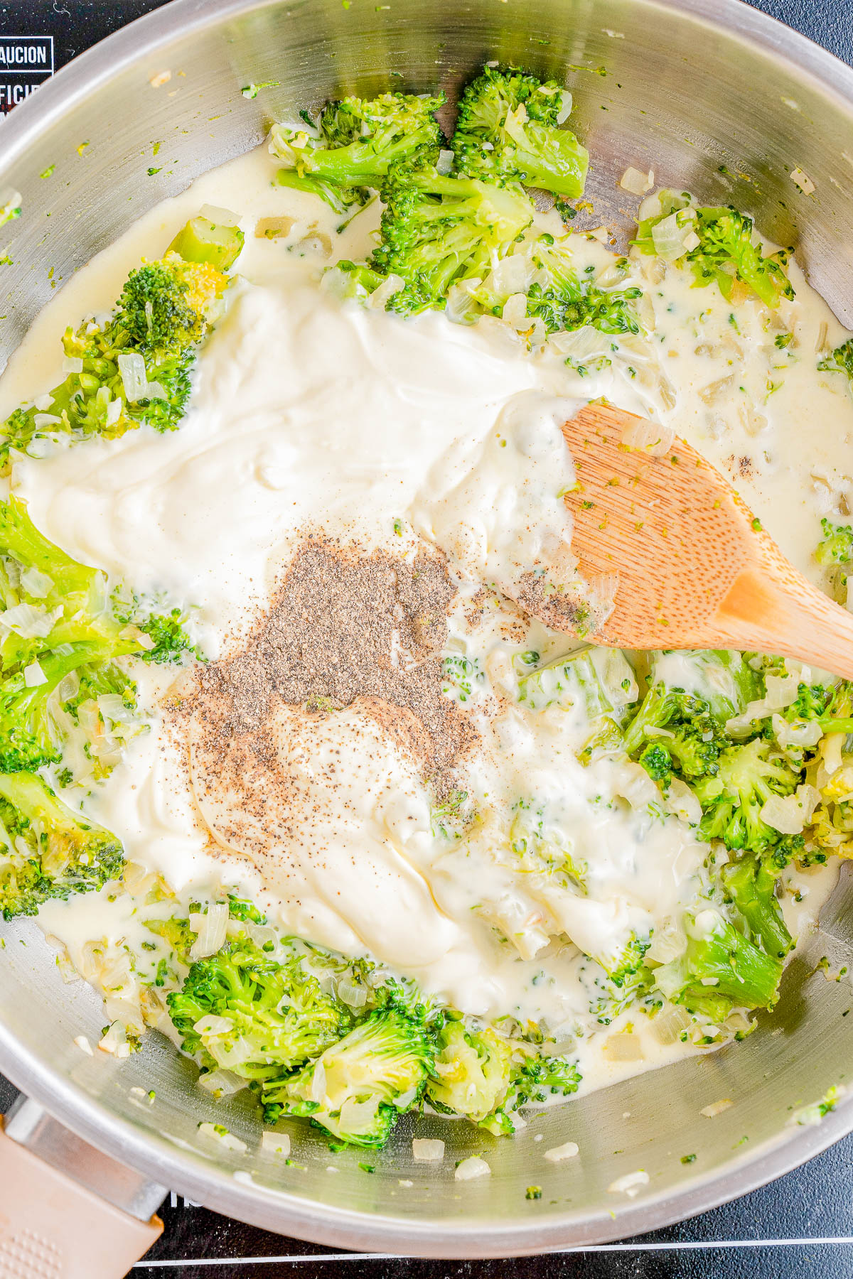 A pan with broccoli, a creamy white sauce, chopped onions, and seasonings being stirred with a wooden spatula.