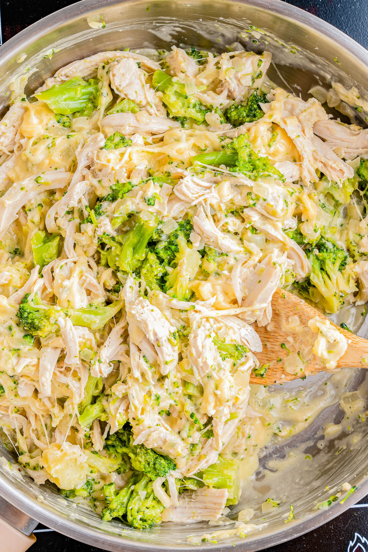 A skillet filled with a creamy chicken and broccoli dish. The mixture includes shredded chicken, pieces of broccoli, and a thick, cheesy sauce. A wooden spoon is partially submerged in the mixture.