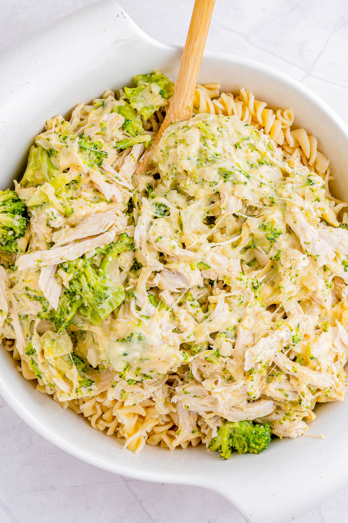 A bowl filled with a mix of cooked fusilli pasta, shredded chicken, broccoli florets, and a creamy sauce. A wooden spoon is placed in the bowl.