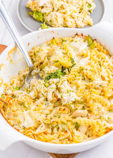 A white casserole dish filled with baked pasta, broccoli, and chicken, served with a spoon. Another portion is plated in the background on a gray plate.