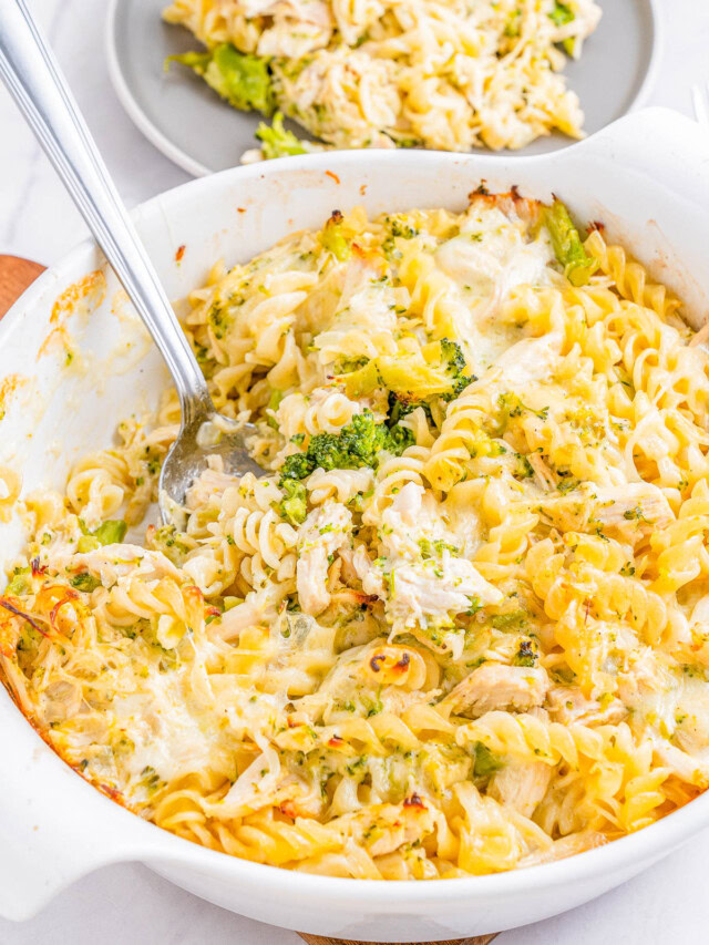 A white casserole dish filled with baked pasta, broccoli, and chicken, served with a spoon. Another portion is plated in the background on a gray plate.