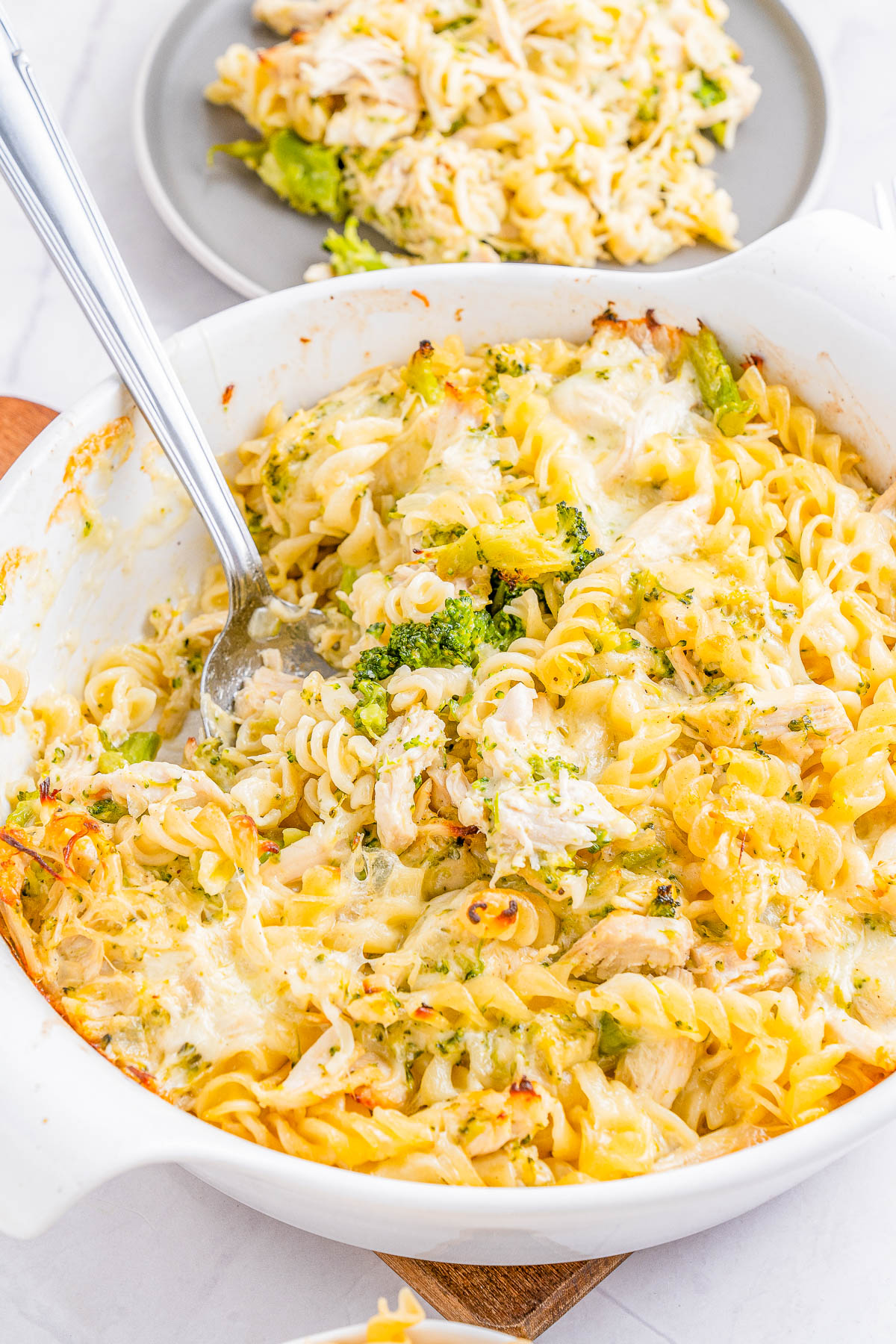 A white casserole dish filled with baked pasta, broccoli, and chicken, served with a spoon. Another portion is plated in the background on a gray plate.