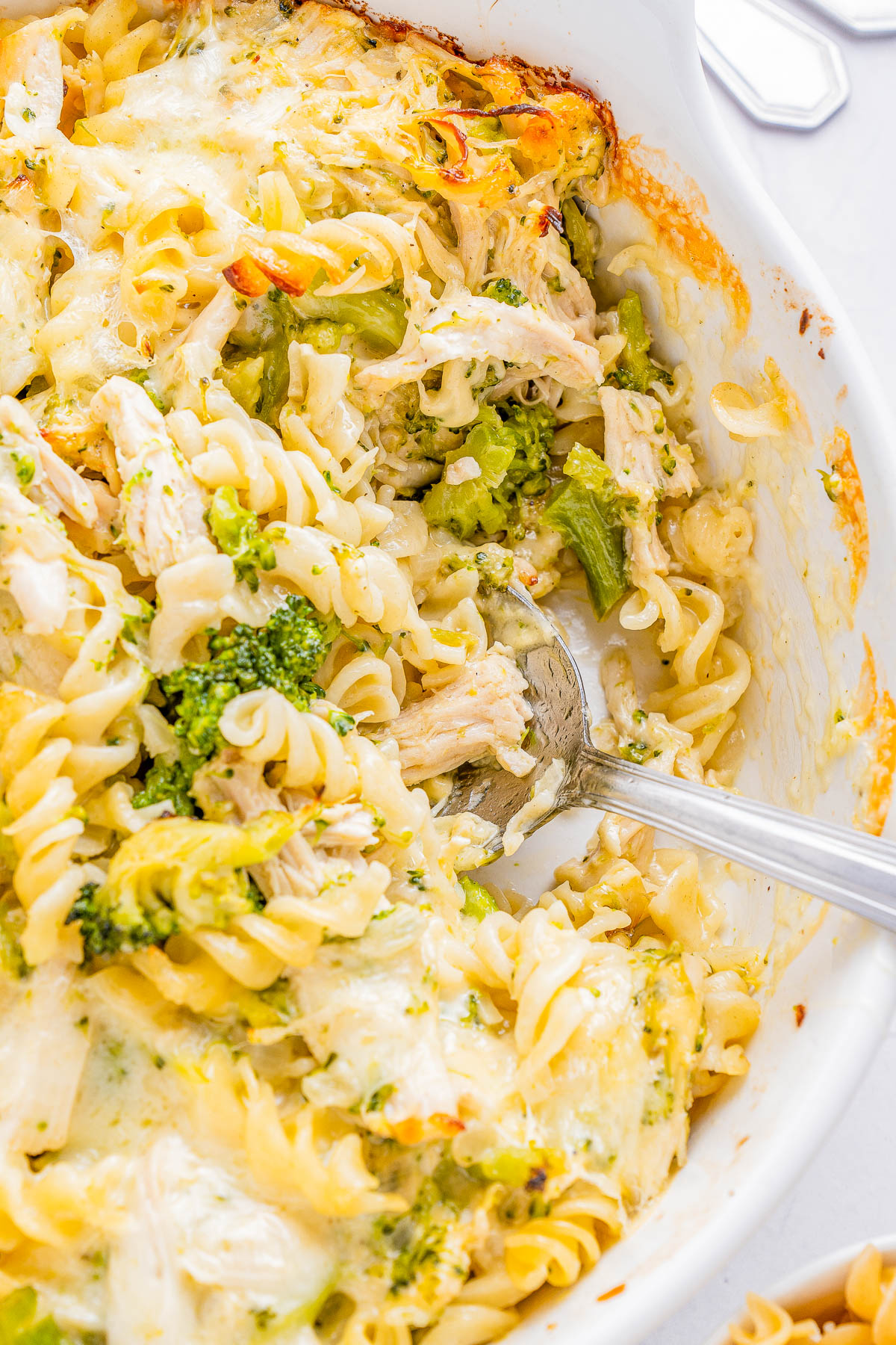 A close-up of a baked pasta dish featuring spiraled noodles, broccoli, and chunks of chicken, with melted cheese on top. A serving spoon is placed in the dish.