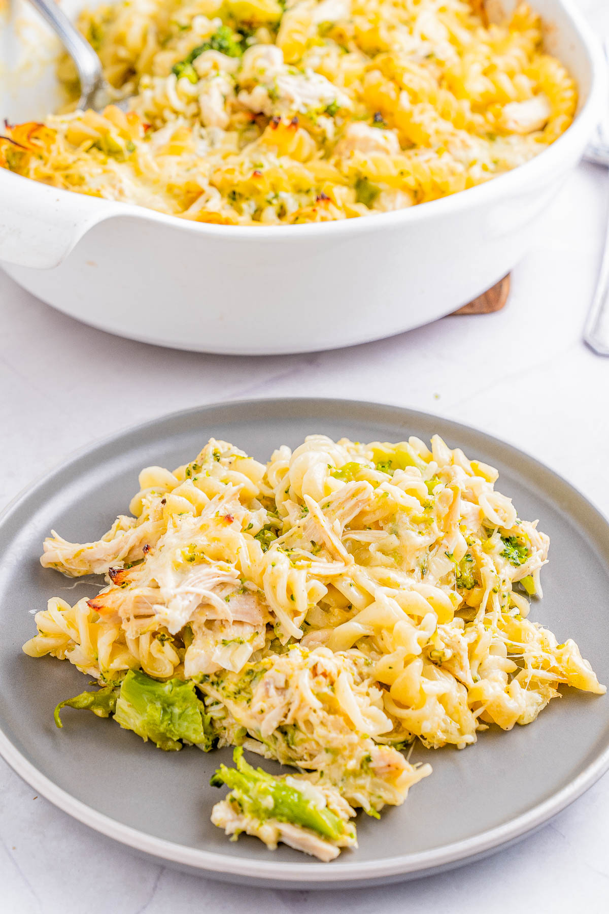 A plate of baked pasta with melted cheese, pieces of chicken, and broccoli is served. A casserole dish of the same pasta is in the background.