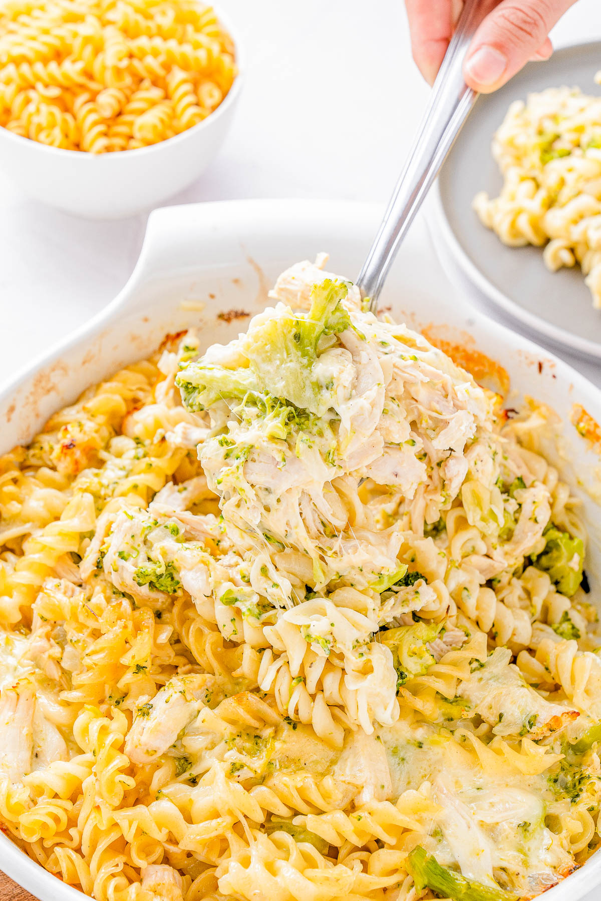 A hand is serving cheesy pasta casserole with broccoli from a white dish, with a bowl of uncooked pasta in the background.