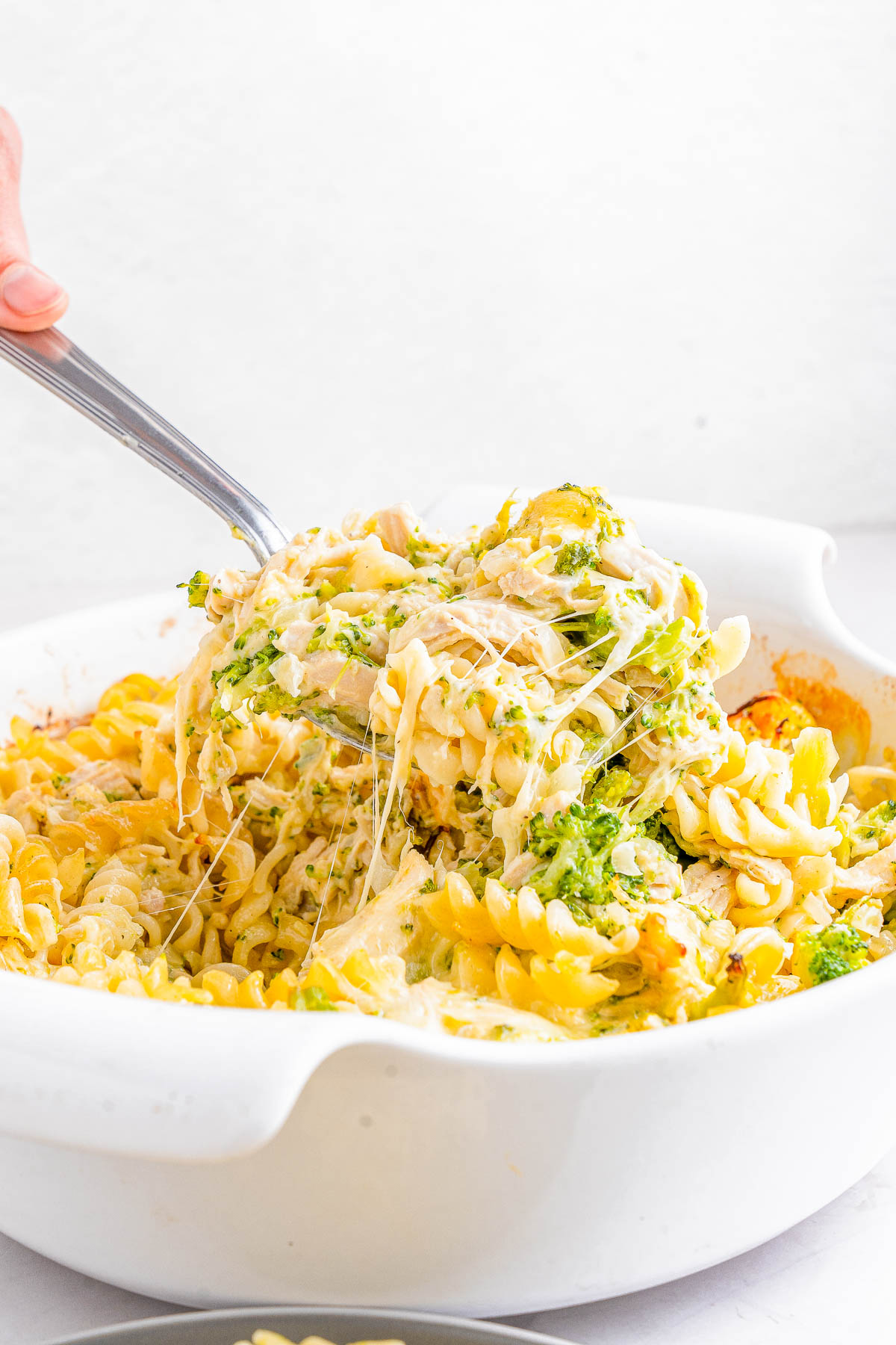 A serving spoon lifts a portion of cheesy broccoli and pasta bake from a white dish.