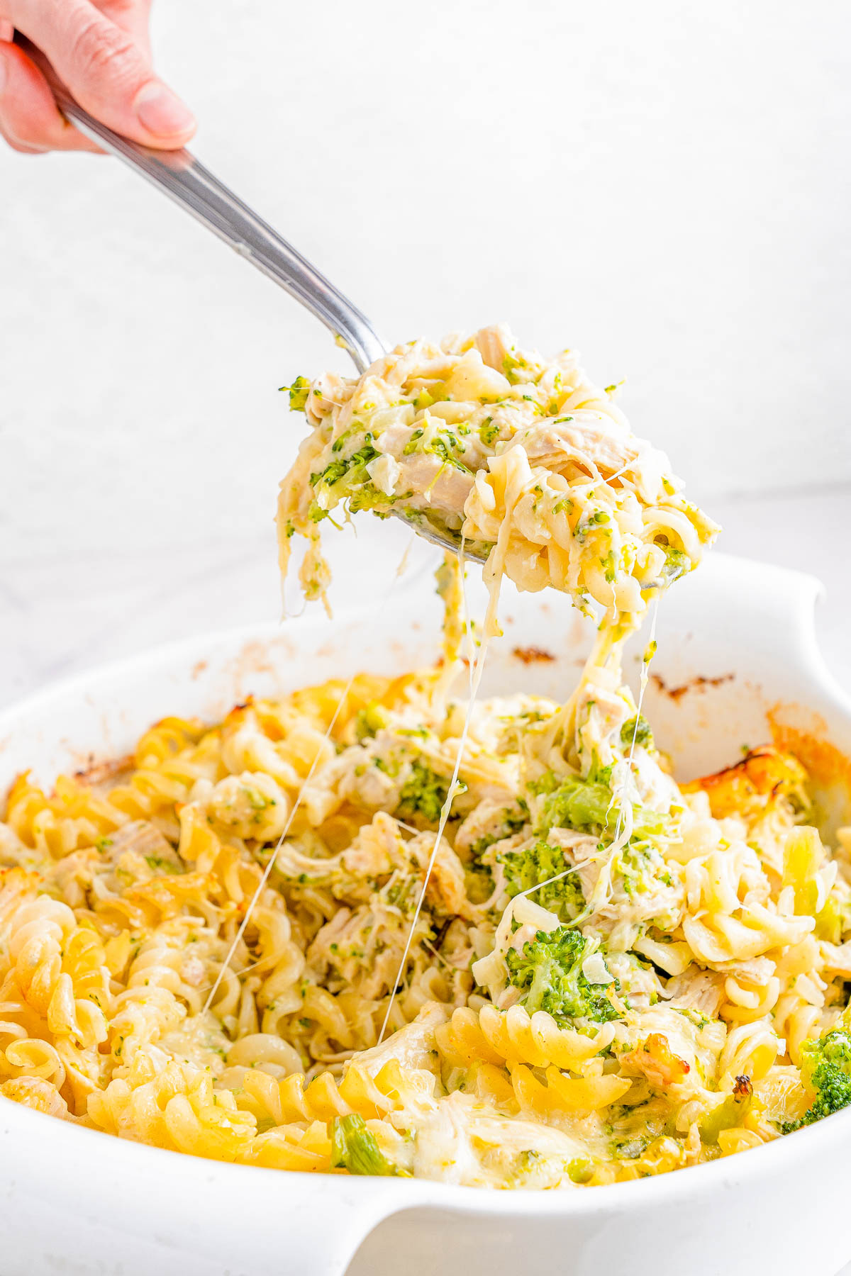 A hand holding a serving utensil scoops out cheesy baked pasta with broccoli from a white casserole dish.