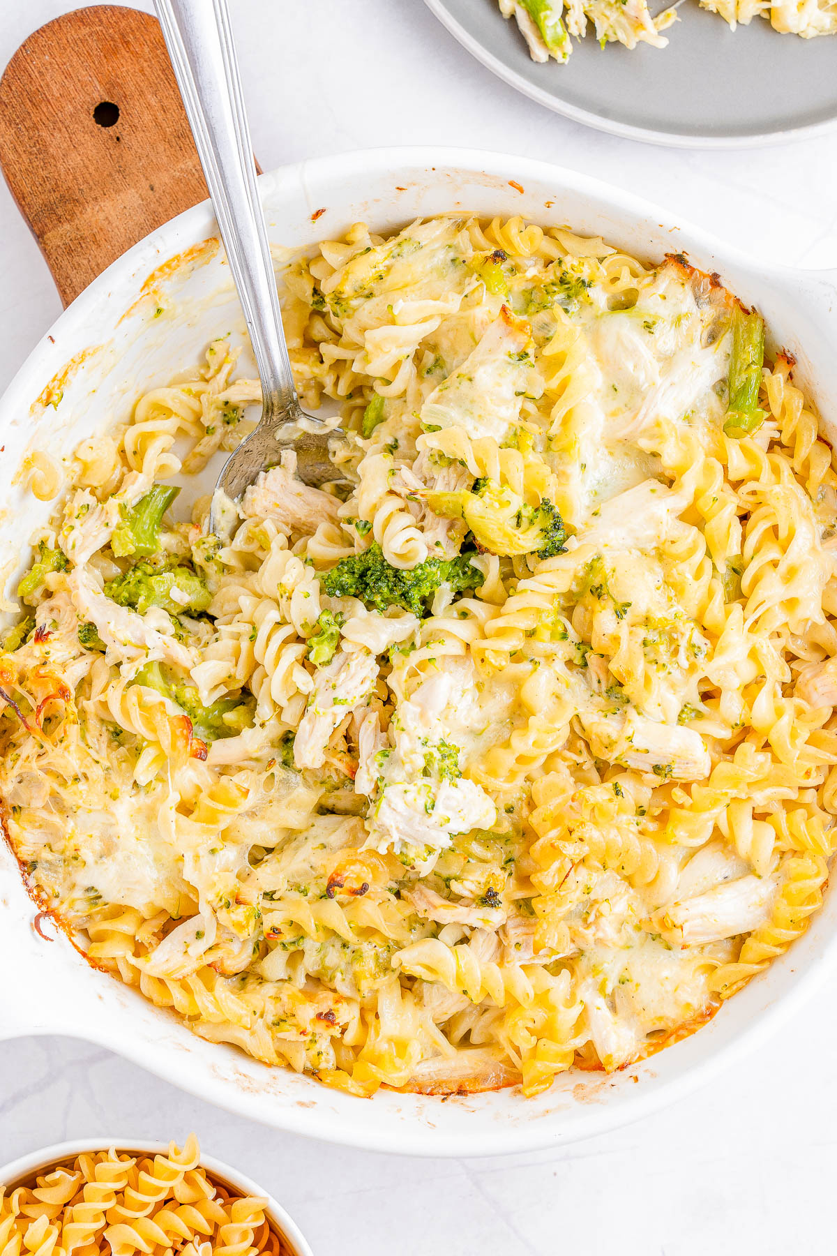 A baked pasta casserole with rotini, chunks of chicken, and broccoli florets in a creamy sauce, displayed in a white casserole dish with a serving spoon.