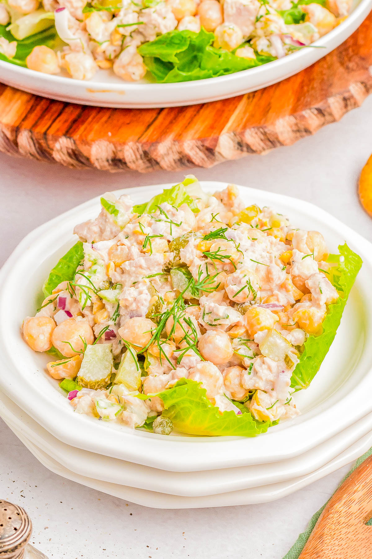 A white plate with a serving of chickpea salad featuring chopped vegetables, lettuce, and a creamy dressing, garnished with fresh dill. Another plate with the same salad is visible in the background.