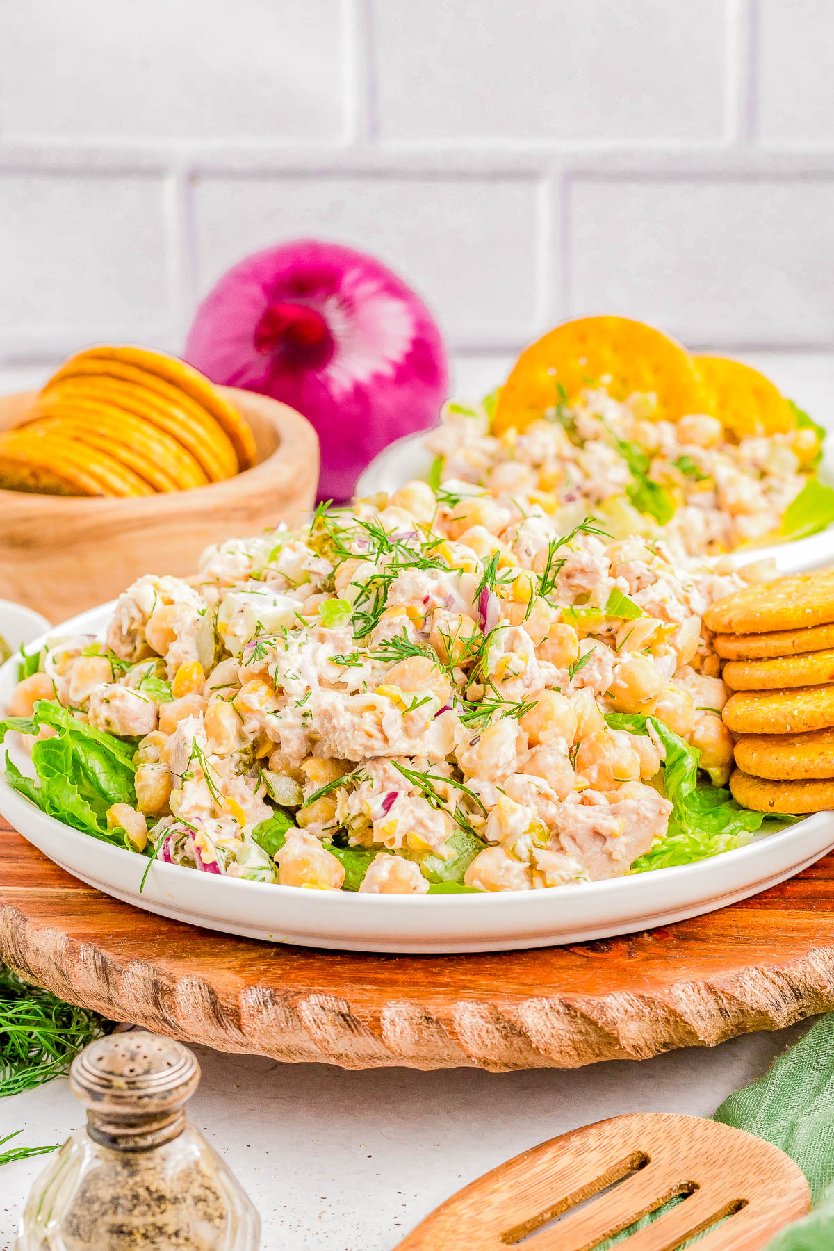 A plate of chickpea salad topped with fresh dill, surrounded by a stack of crackers, a whole red onion, and a wooden bowl filled with additional crackers.