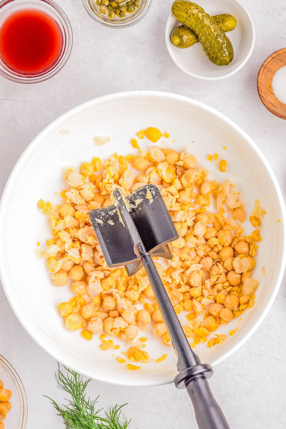 A white bowl contains mashed chickpeas with an avocado masher placed in it. Surrounding the bowl are small dishes with ingredients including pickles, red liquid, capers, and dill.