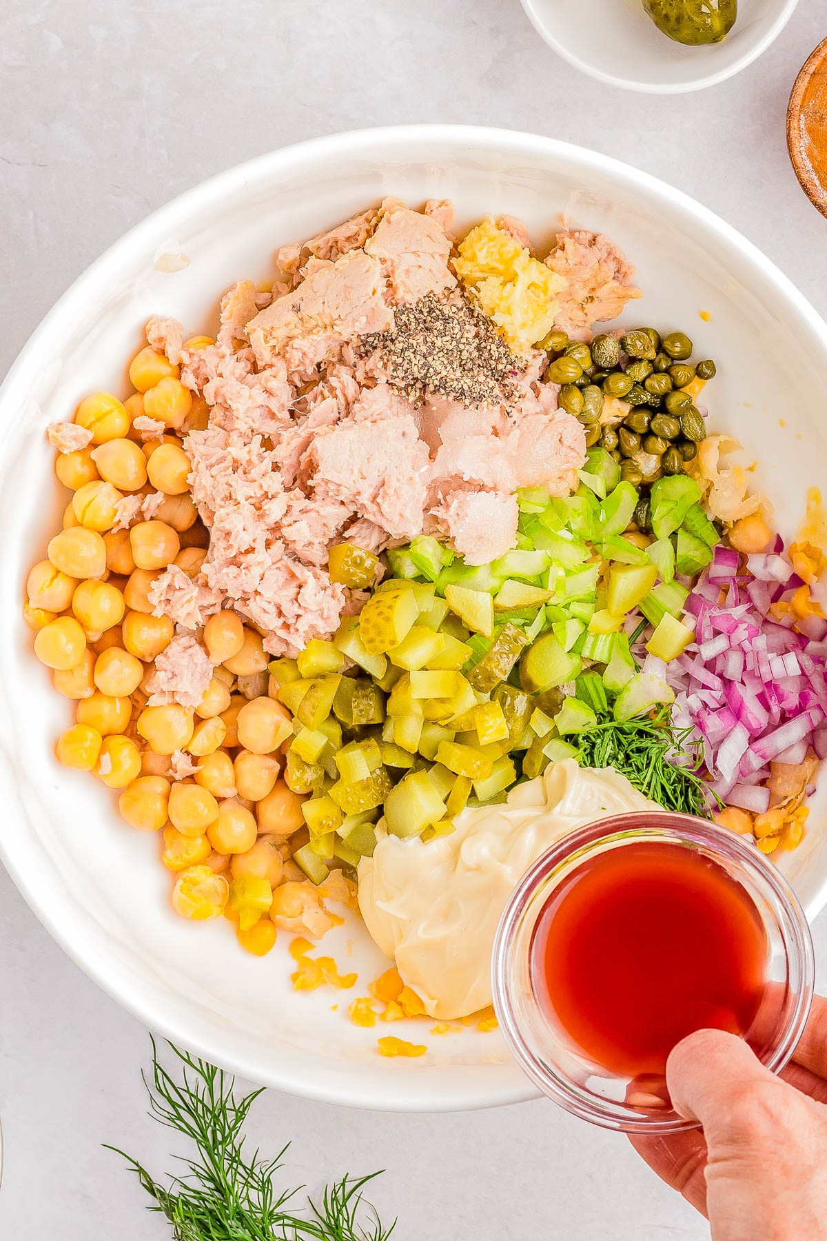 A white bowl containing chickpeas, diced celery, pickles, tuna, red onions, capers, mayonnaise, and chopped dill. A hand is holding a small glass bowl of red liquid, about to pour it into the mix.