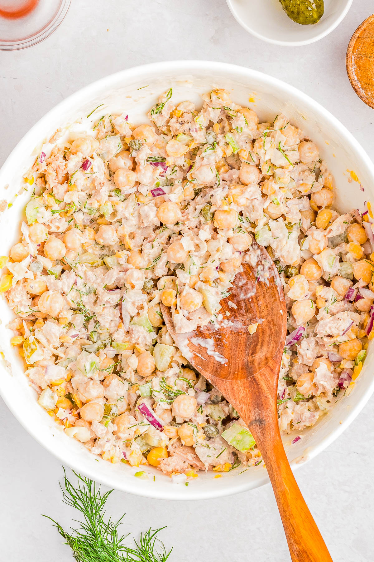 A bowl filled with a chickpea salad mixture, featuring chickpeas, chopped red onions, and other vegetables, with a wooden spoon resting inside. Fresh dill is placed nearby on a light-colored surface.