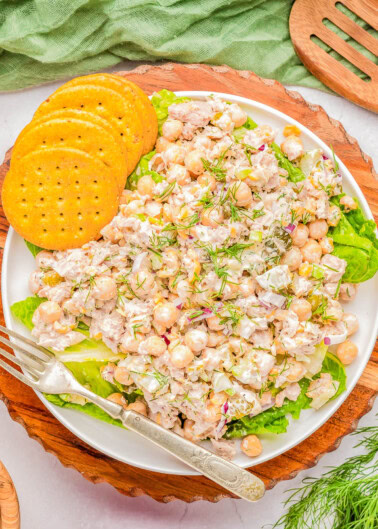 A plate of chickpea salad with visible ingredients including chickpeas, herbs, and dressing, garnished with fresh dill. The plate is accompanied by three round crackers and a fork, placed on a wooden board.