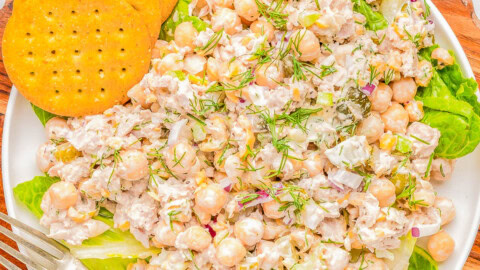 A plate of chickpea salad with visible ingredients including chickpeas, herbs, and dressing, garnished with fresh dill. The plate is accompanied by three round crackers and a fork, placed on a wooden board.