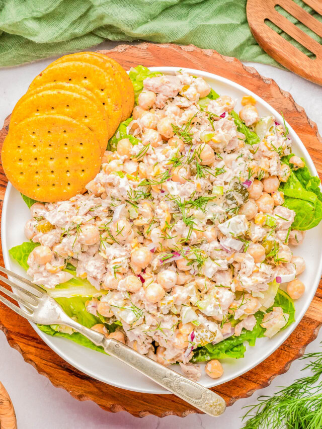 A plate of chickpea salad with visible ingredients including chickpeas, herbs, and dressing, garnished with fresh dill. The plate is accompanied by three round crackers and a fork, placed on a wooden board.