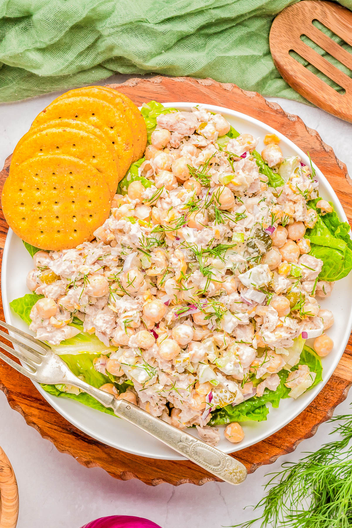 A plate of chickpea salad with visible ingredients including chickpeas, herbs, and dressing, garnished with fresh dill. The plate is accompanied by three round crackers and a fork, placed on a wooden board.