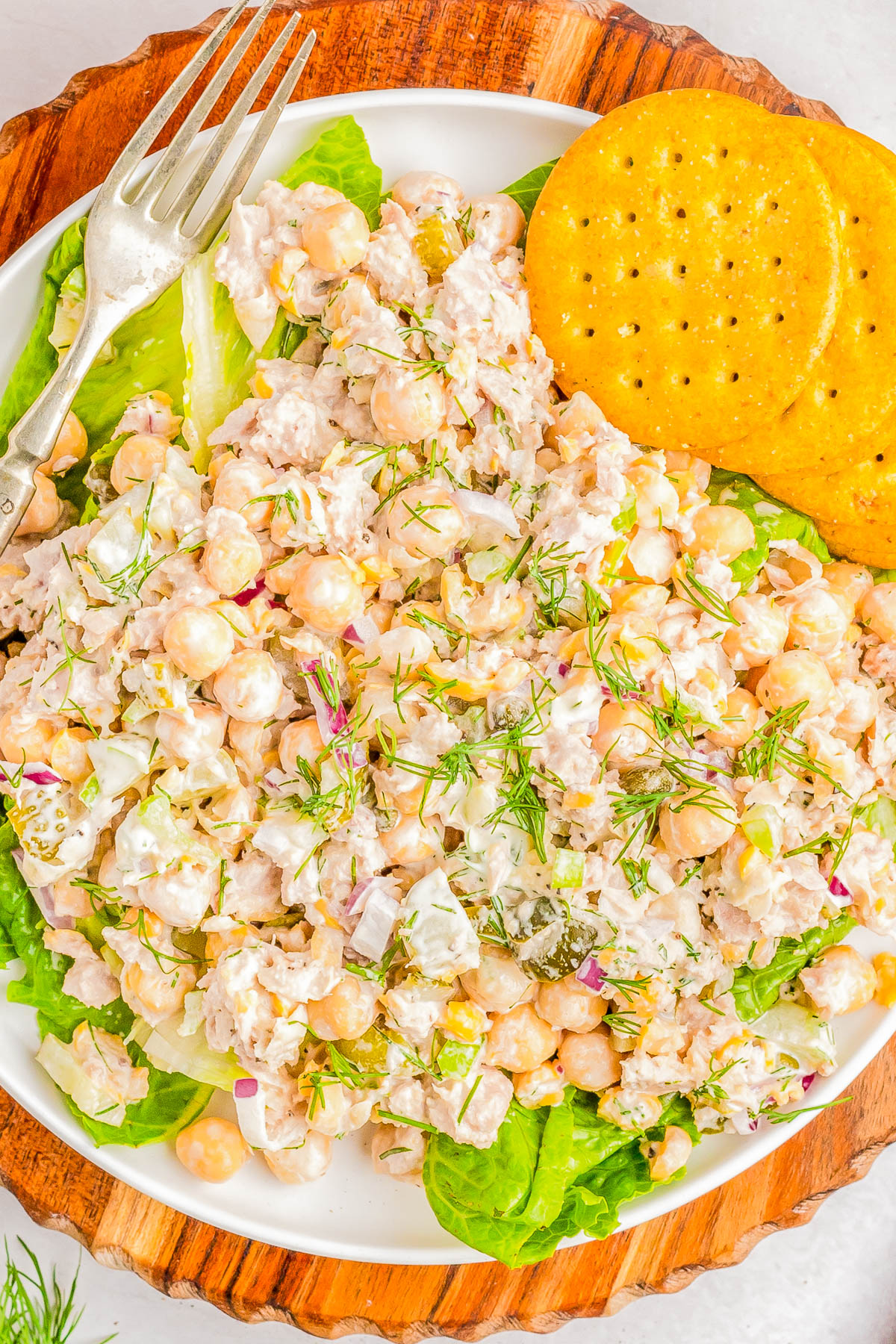 A white plate contains a salad made with chickpeas, diced onions, and herbs, served on a bed of lettuce leaves. Two round crackers and a fork are placed on the side of the plate.