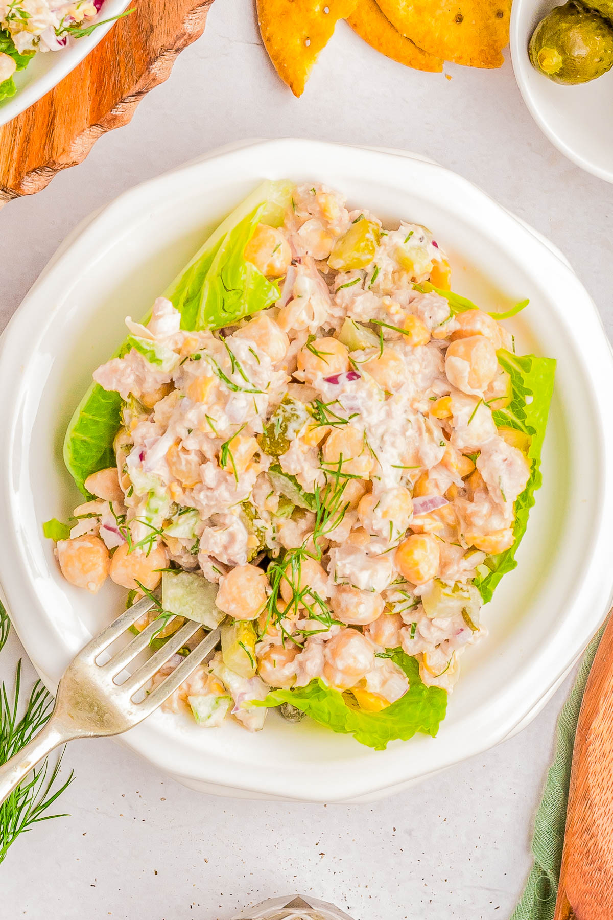 A white plate with a chickpea salad garnished with fresh dill and served on lettuce leaves, accompanied by a fork.