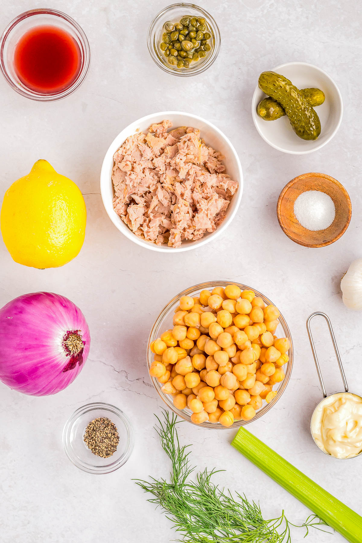 Ingredients laid out on a counter include chickpeas, tuna, red onion, lemon, celery, fresh dill, pickles, capers, black pepper, salt, red wine vinegar, garlic, and mayonnaise.