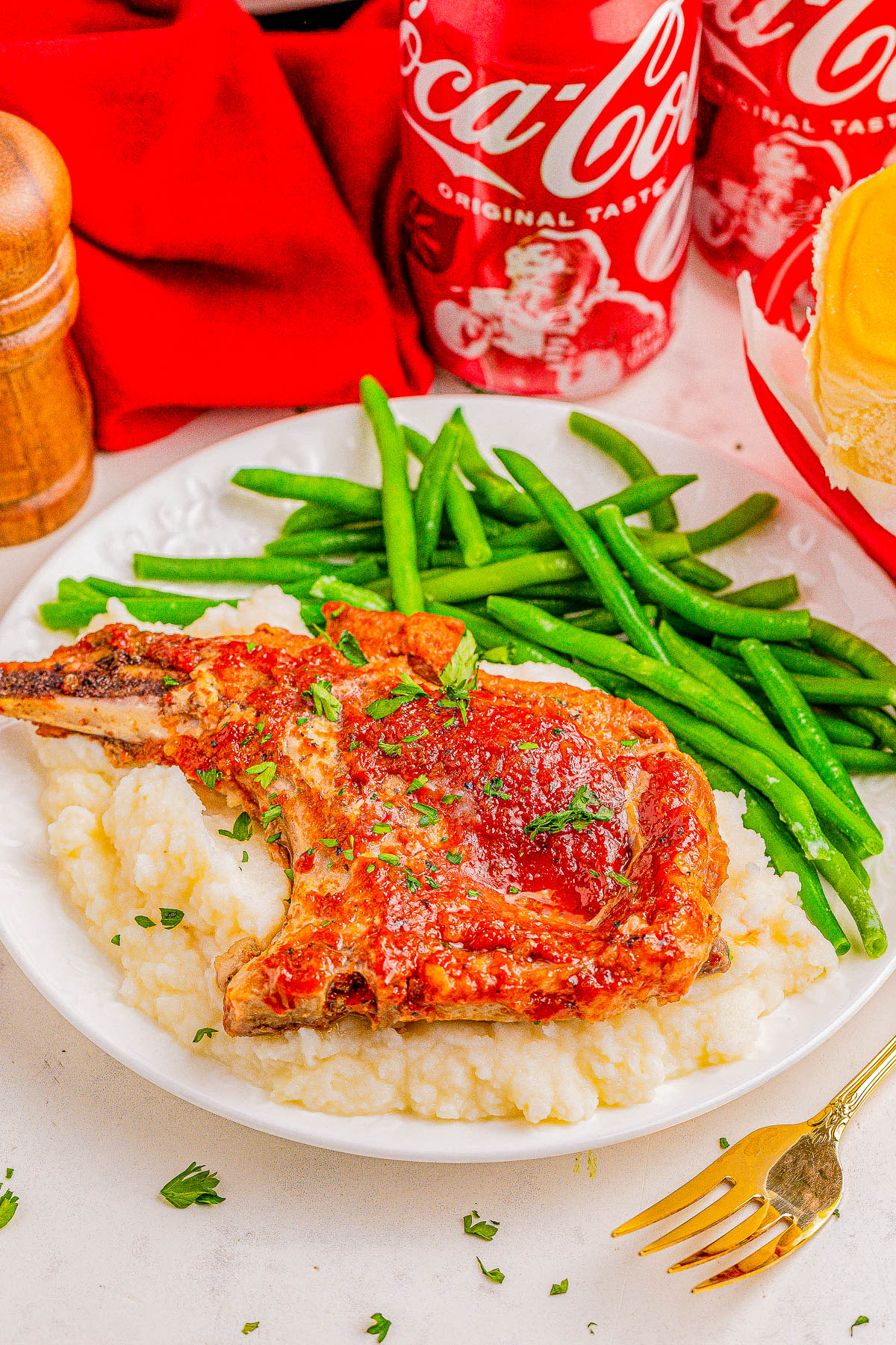 A plate with a pork chop covered in sauce, served on mashed potatoes with green beans on the side. Two cans of Coca-Cola are in the background.