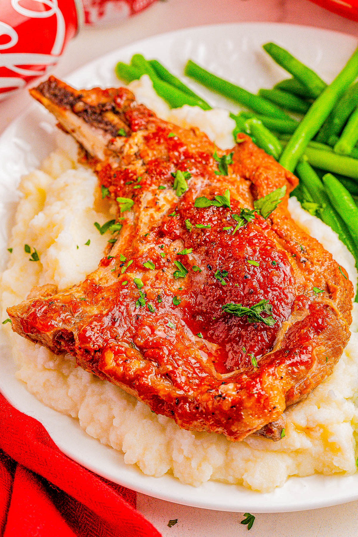 A plate with a crispy pork chop topped with sauce, served on a bed of mashed potatoes, accompanied by a side of green beans.