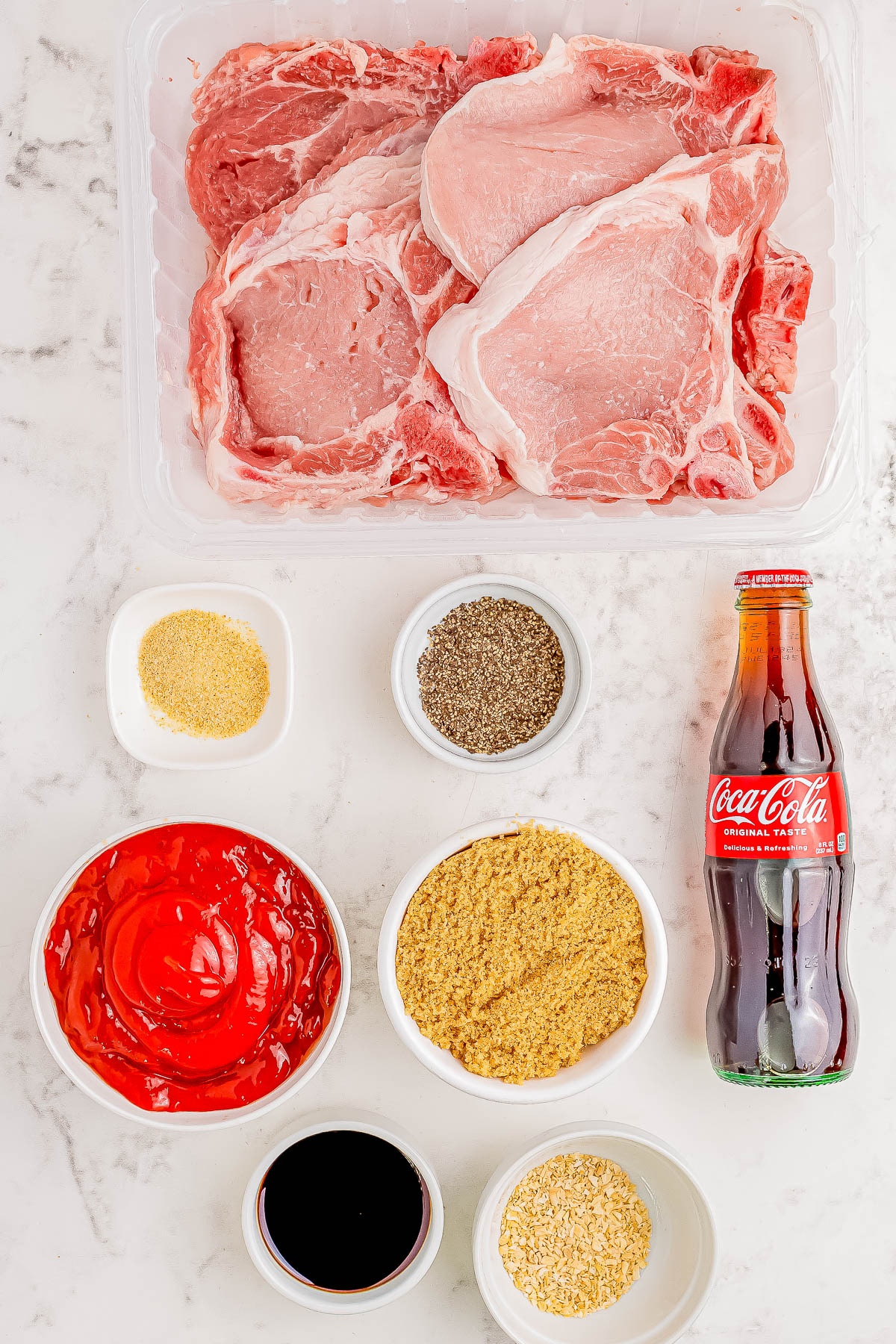 Raw pork chops on a tray surrounded by spices, ketchup, a small bowl of soy sauce, mustard, and a bottle of Coca-Cola on a marble surface.