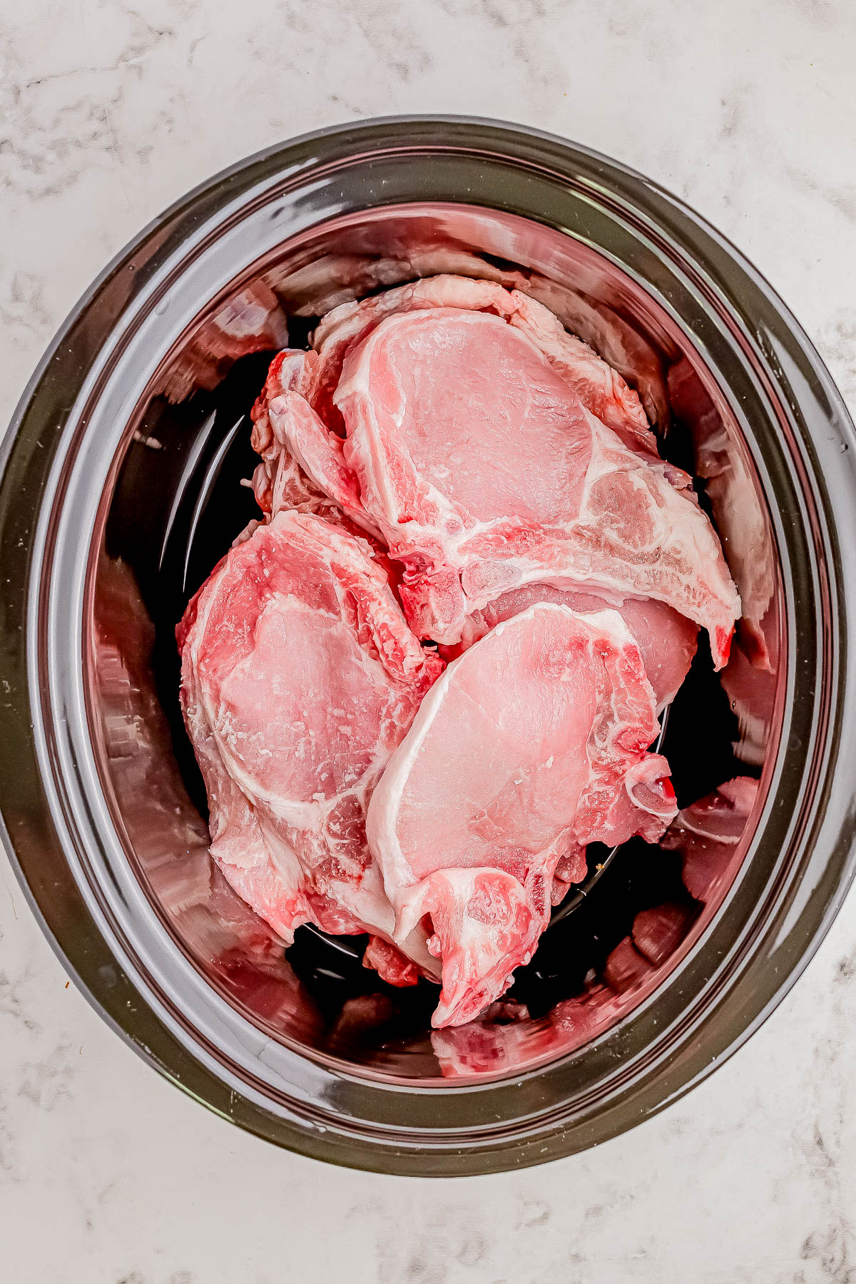 A black slow cooker filled with raw pork chops, placed on a white countertop.