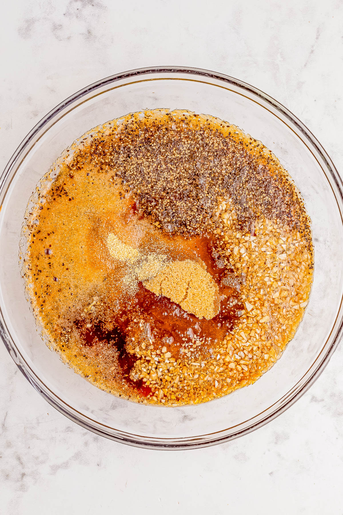 A glass bowl containing a mixture of various spices and liquids, including chopped garlic, black pepper, mustard powder, and other seasonings on a light-colored surface.