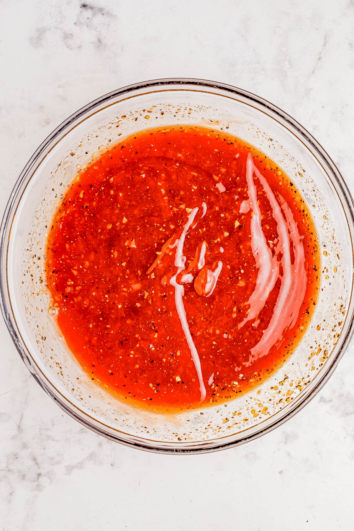 A clear glass bowl filled with red, chunky liquid that appears to be salsa or a sauce, placed on a white marble surface.