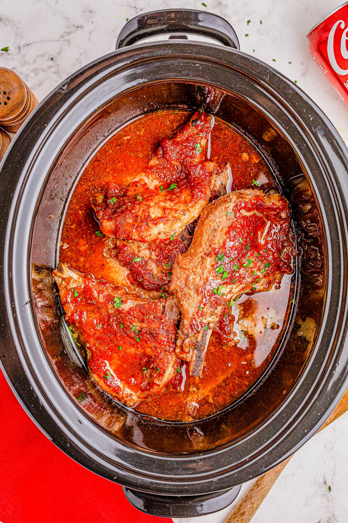 A slow cooker with cooked meat in a rich, red sauce garnished with parsley, positioned on a marble counter next to a can of Coca-Cola and wooden salt and pepper shakers.