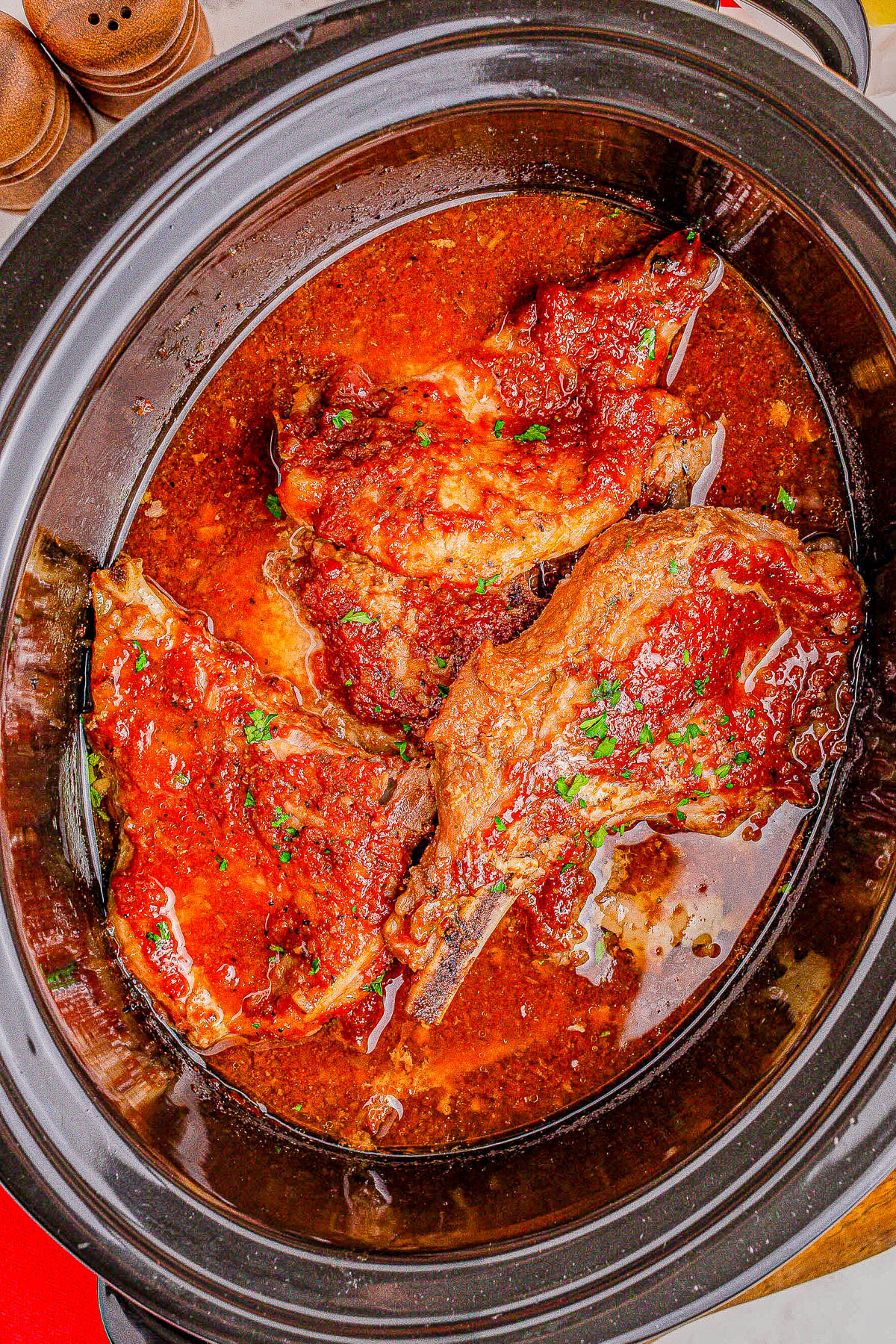 Close-up of three cooked pork chops in a crockpot, immersed in a red tomato-based sauce and garnished with chopped herbs.