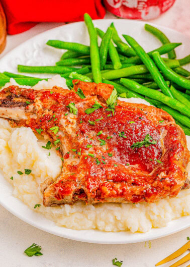 A plate with a pork chop garnished with herbs on a bed of mashed potatoes, accompanied by green beans.