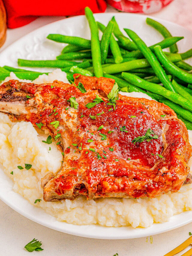 A plate with a pork chop garnished with herbs on a bed of mashed potatoes, accompanied by green beans.