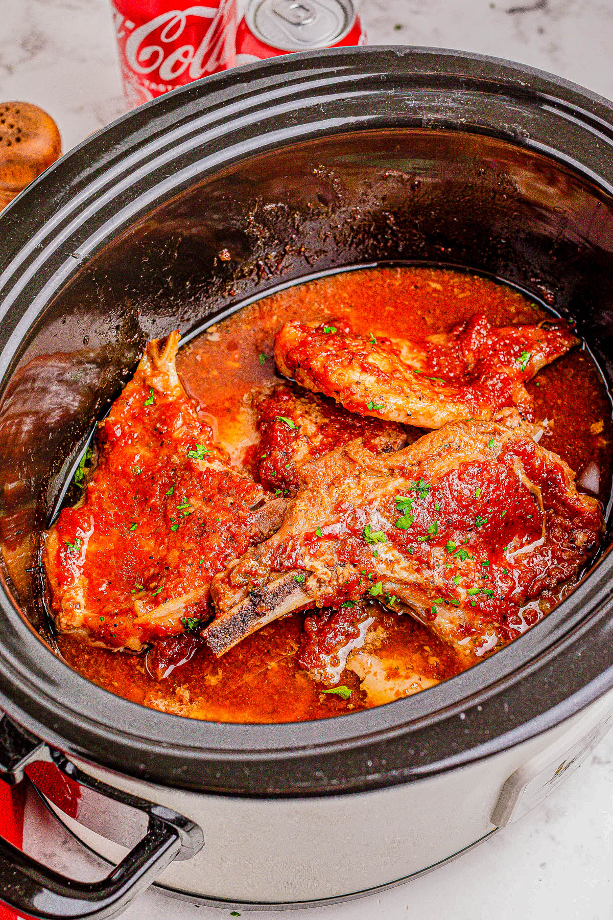 A slow cooker filled with cooked pork chops in a red sauce. A napkin and two opened cans of Coca-Cola are visible in the background.