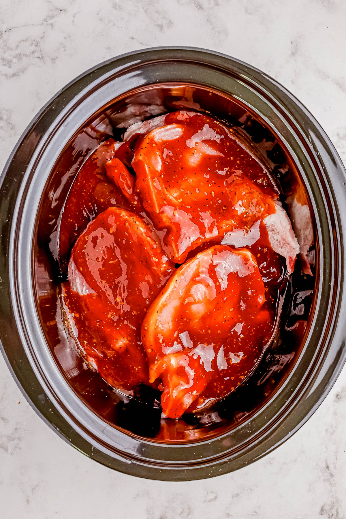 Raw chicken breasts marinated in a sauce, placed inside a crockpot on a marble surface.