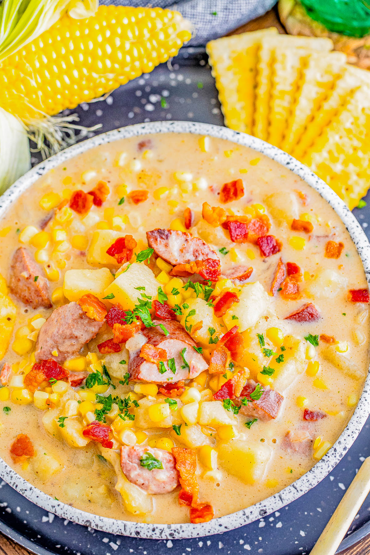 A bowl of creamy corn chowder with pieces of sausage, bacon bits, and chopped herbs on top. Corn on the cob and crackers are in the background.